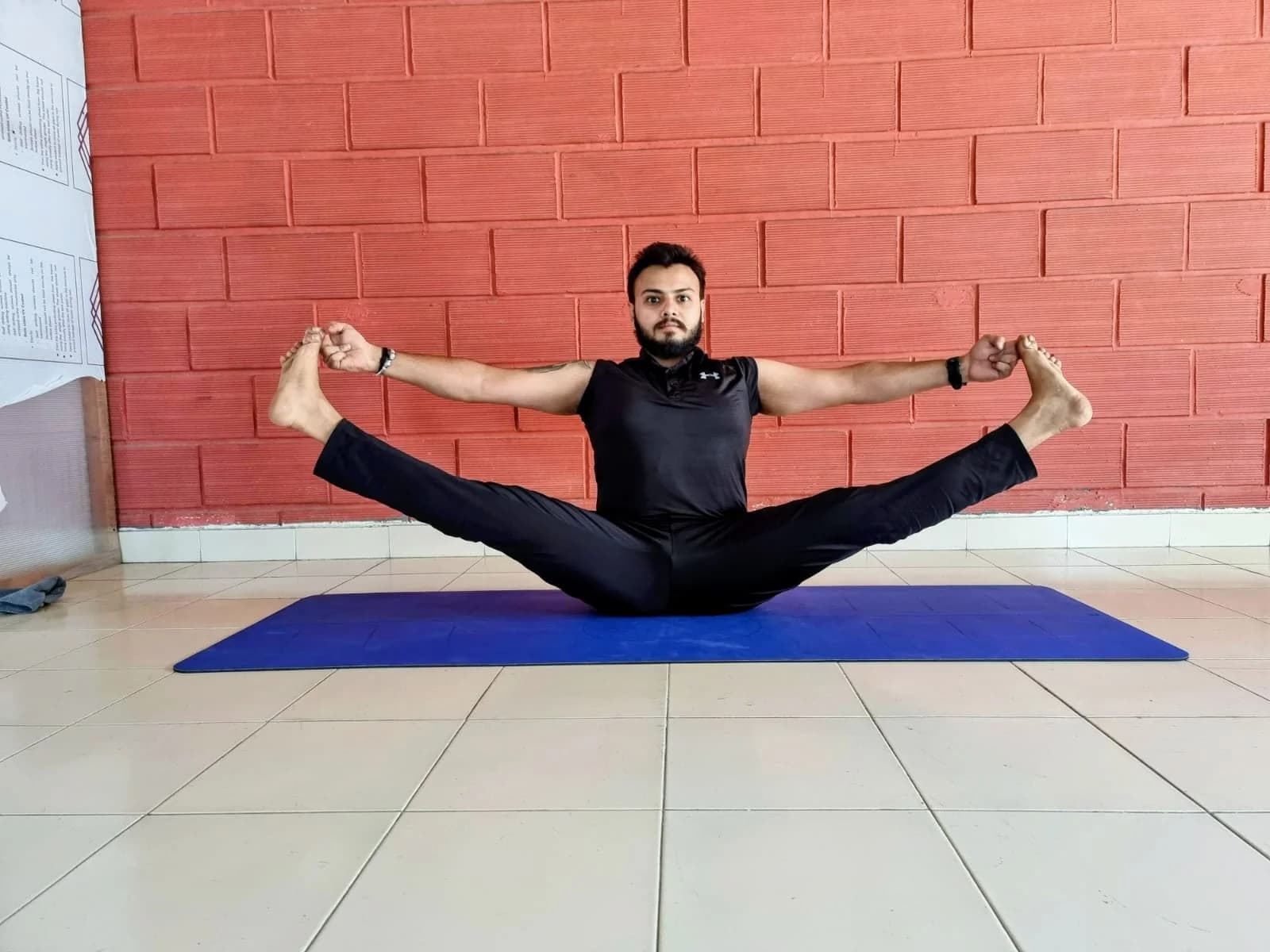 Gandharva in a wide-legged toe-hold forward fold against a brick wall