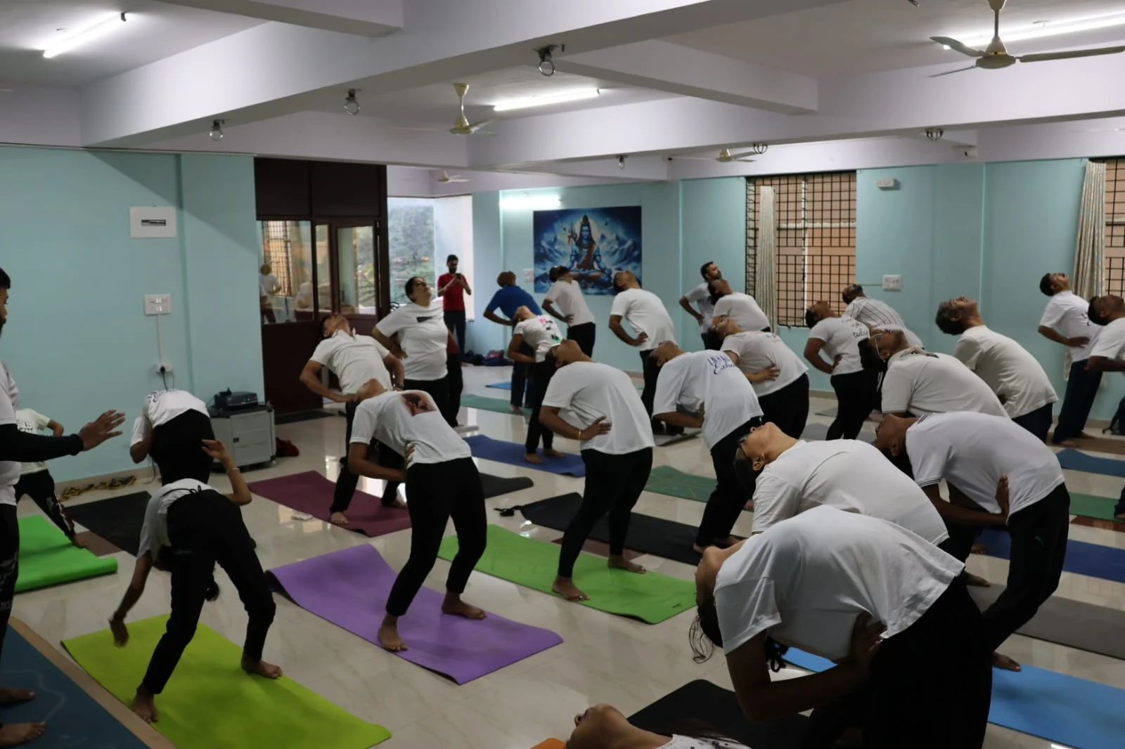 A full class in a synchronized backbend row, framed by a window view