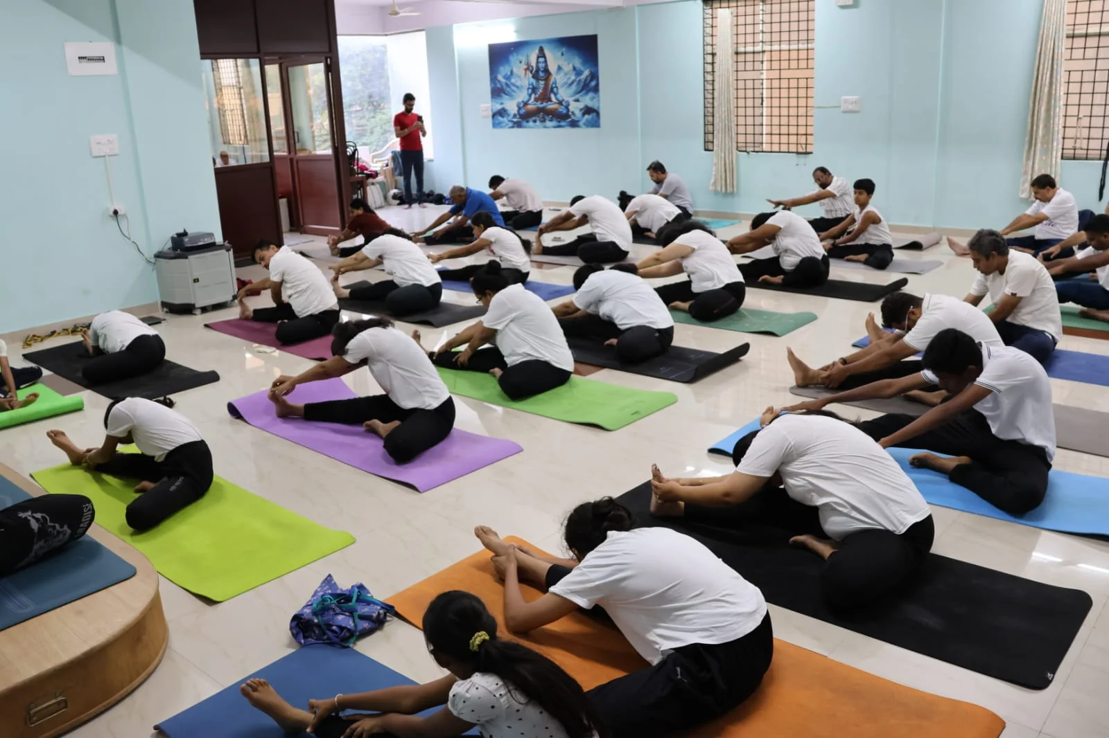 A full class in a seated forward-fold, colorful mats lining the floor