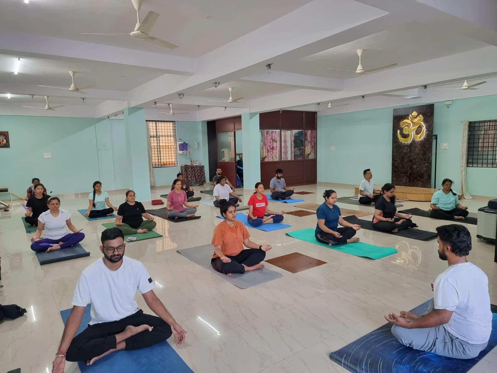 A large group seated in meditation in symmetric rows beneath a gold Om backdrop