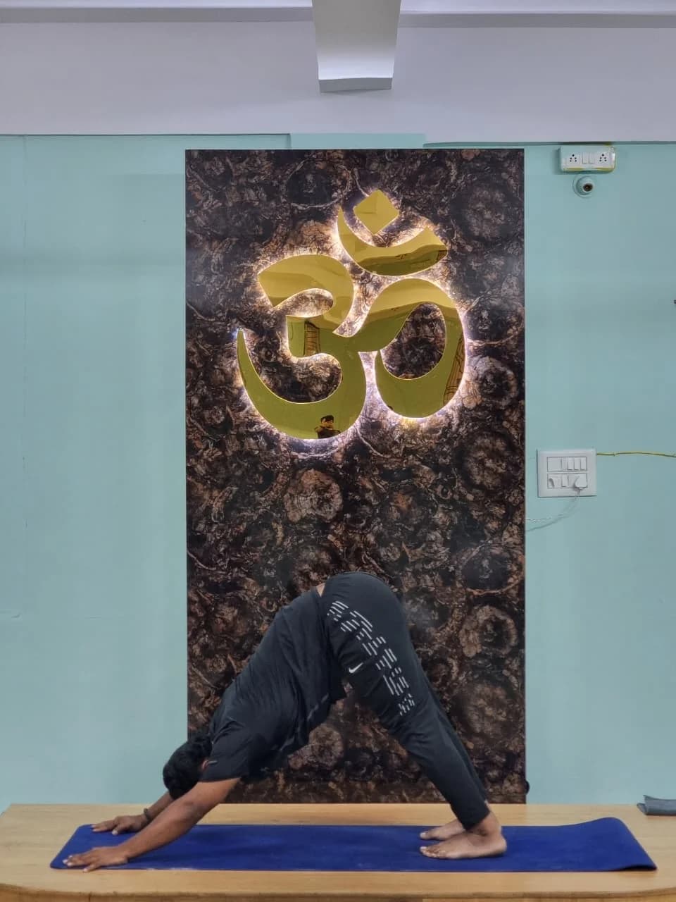 A solo practitioner in downward-dog directly beneath a backlit gold Om sign
